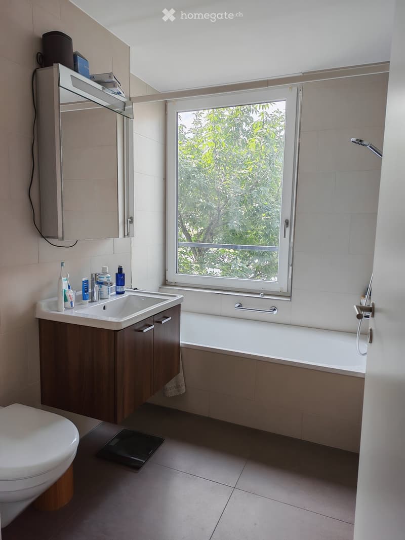 Modern bathroom with a large window, a white bathtub, a dark wooden sink cabinet, a white toilet, and a mirror above the sink. Various toiletries are placed on the sink counter.
