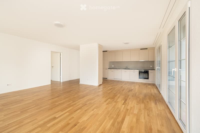 Open-plan unfurnished room with light wooden flooring, white walls, and a small kitchen area with cabinets and appliances.