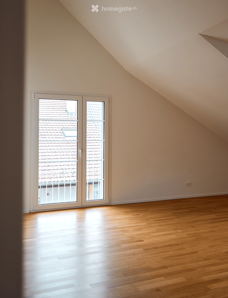 Empty attic room with wooden flooring, large window showing a roof view, and slanted ceilings.