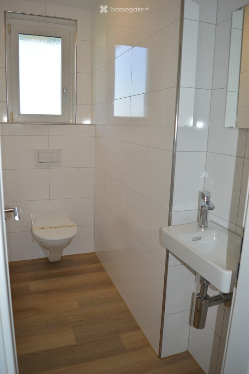 Modern bathroom with white tiled walls, wooden floor, toilet, sink, and chrome fixtures.