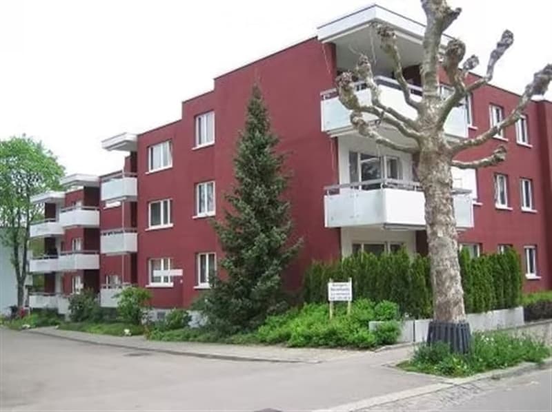 Red apartment building with multiple balconies and white trim, surrounded by greenery and trees.