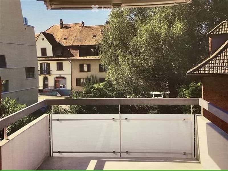 Balcony with metal railing and sliding glass doors, overlooking a tree-lined street and neighboring buildings.