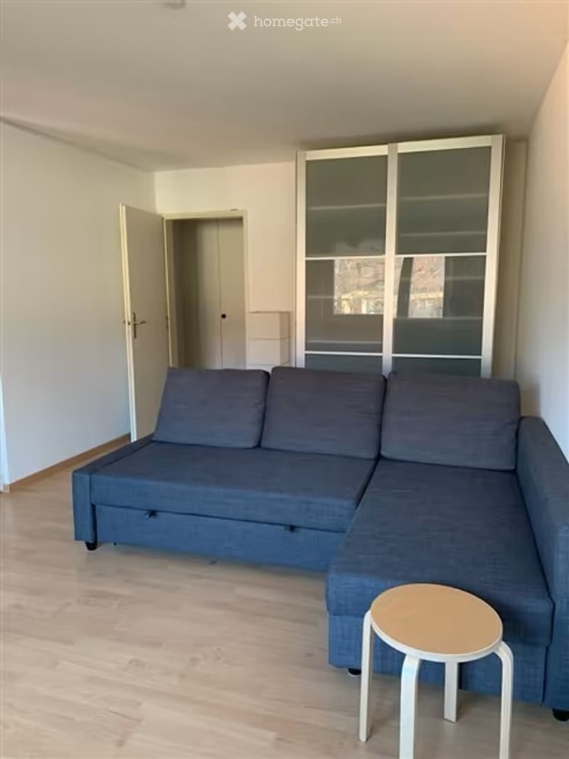A modern living room with a grey sectional sofa and a small round stool, featuring light wooden flooring and white walls.