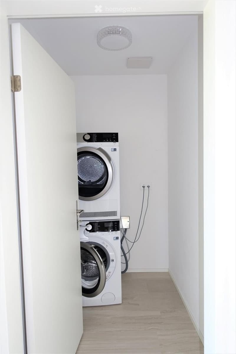A small, modern bathroom with a washing machine and dryer unit, white walls, and wooden flooring.