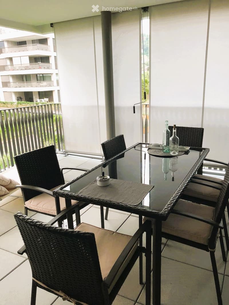 Balcony with a glass table, wicker chairs, and two bottles on the table. The balcony has a modern railing and overlooks another building.