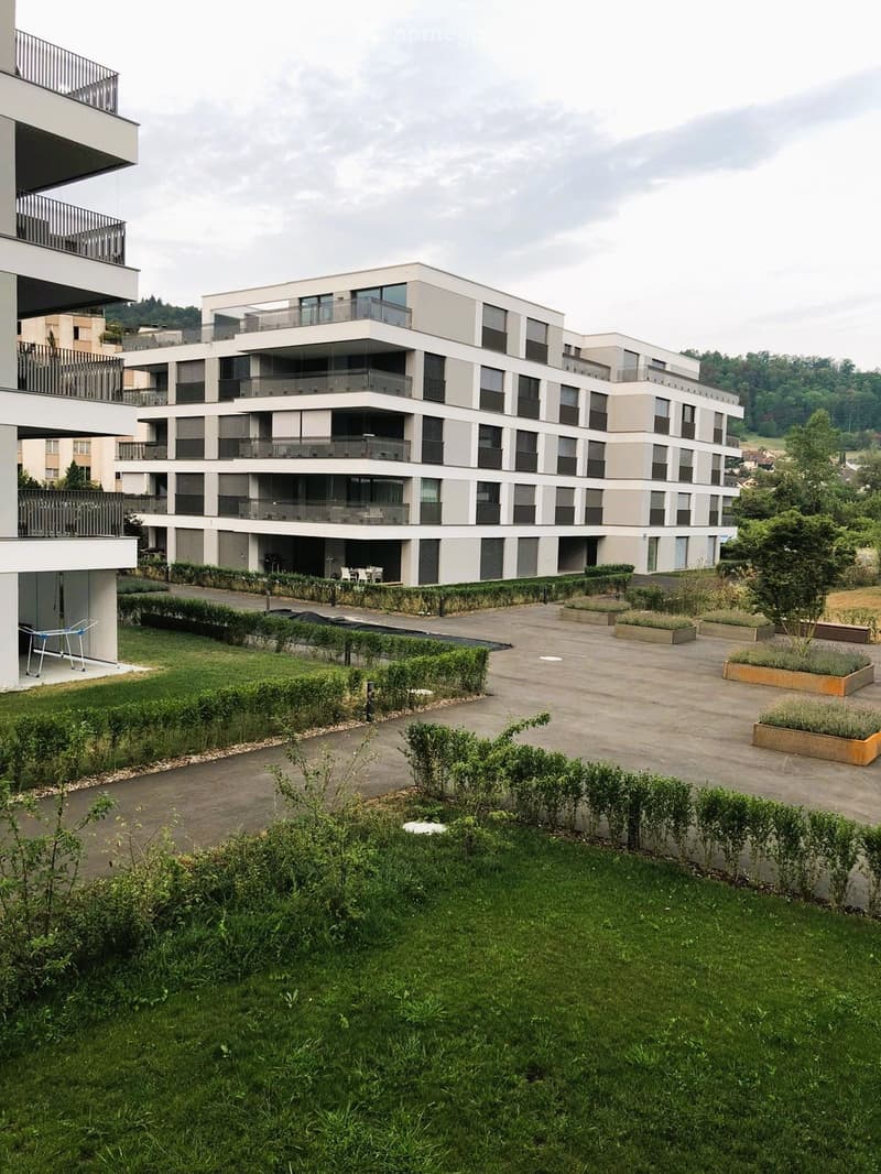 Modern multi-story residential building with balconies, surrounded by landscaped gardens and pathways.