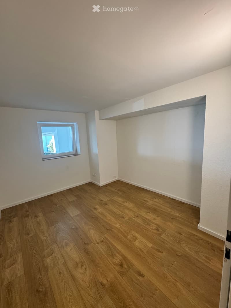 Empty room with wooden flooring, white walls, and a single window with a view of greenery outside.