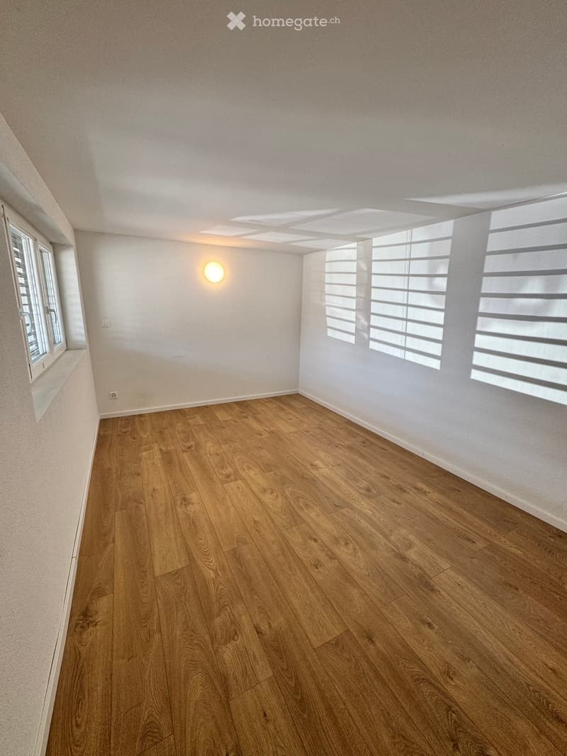 Empty room with wooden flooring, white walls, and large windows with blinds. A single wall-mounted light fixture is visible.
