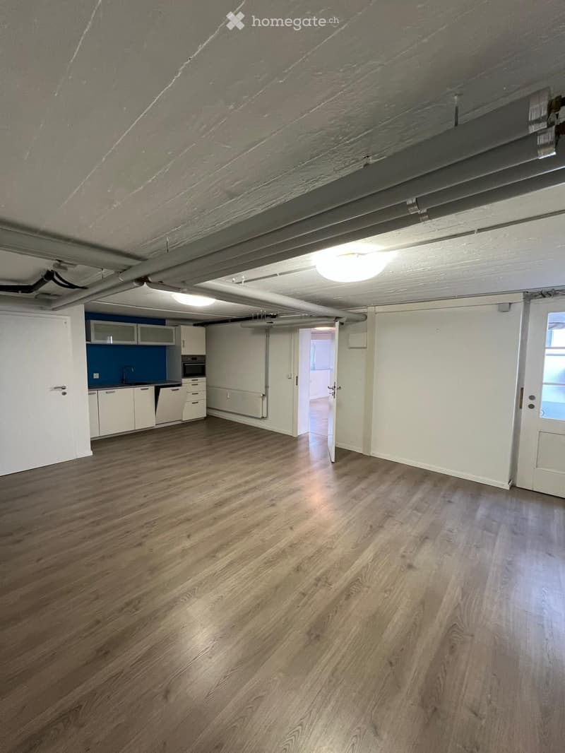 Empty room with wooden flooring, white walls, and exposed ceiling beams. A small kitchen area is visible in the background with white cabinets and a blue backsplash.