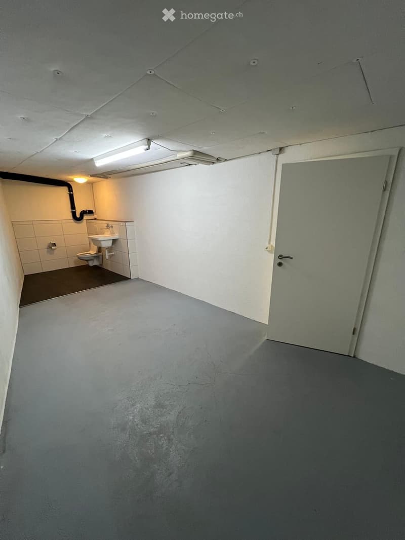 Empty room with grey walls and floor, a closed door, and a small adjacent bathroom with a toilet, sink, and tiled walls.