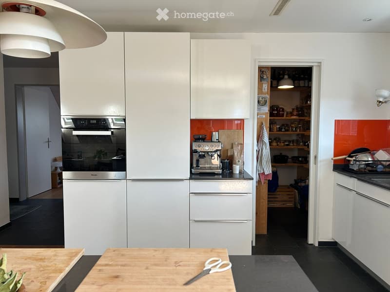 Modern kitchen with white cabinets, stainless steel appliances, and a bright orange backsplash. Features include a refrigerator, oven, coffee machine, and open shelving with jars and bottles.