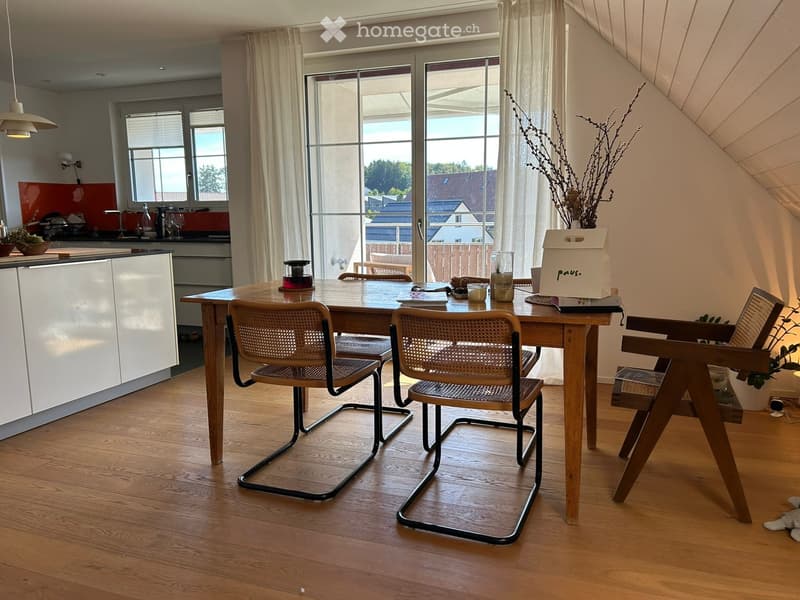A modern dining area with a wooden dining table, four chairs, and a kitchen area visible in the background. The space features large windows with white curtains, wooden flooring, and a sleek kitchen cabinet.