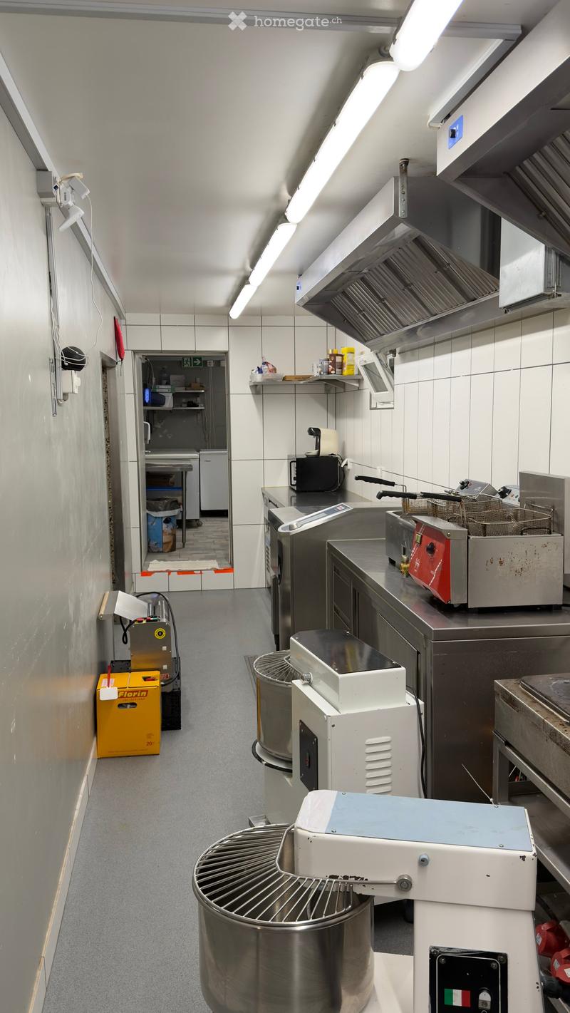 Commercial kitchen with stainless steel appliances, including a large mixer, fryer, oven, and refrigerator. Shelves and a counter are visible, along with an oven hood and various kitchen equipment.