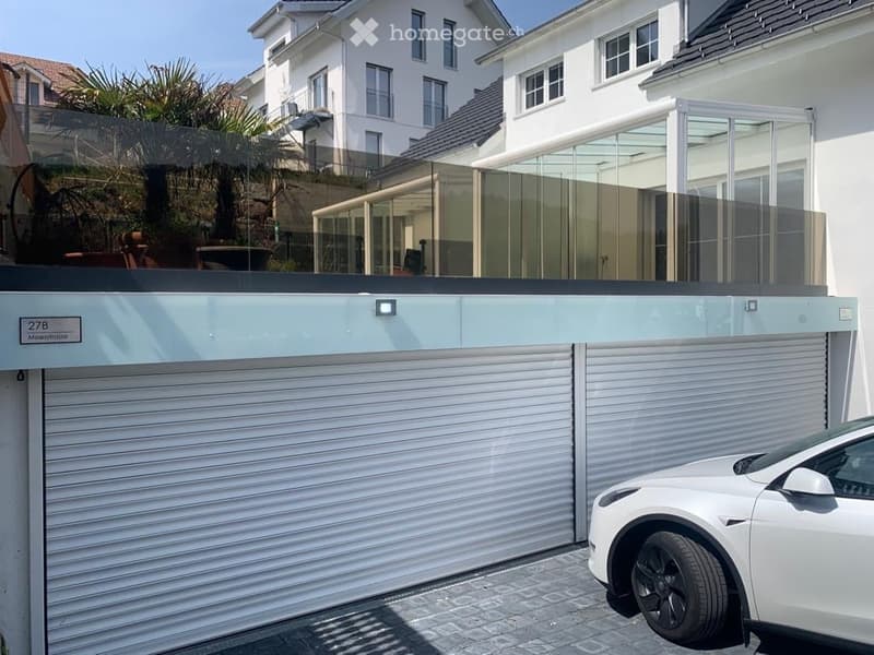 Modern two-story house with a large garage and a sleek, contemporary design. A white car is parked in front of the garage, and the building features large windows and a clean, minimalist aesthetic.