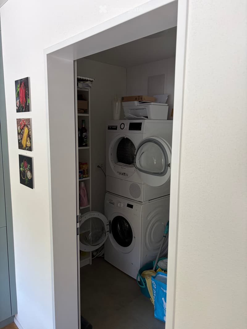 Compact laundry area with a stacked washing machine and dryer, a small shelf with cleaning supplies, and a broom and dustpan in a bucket.