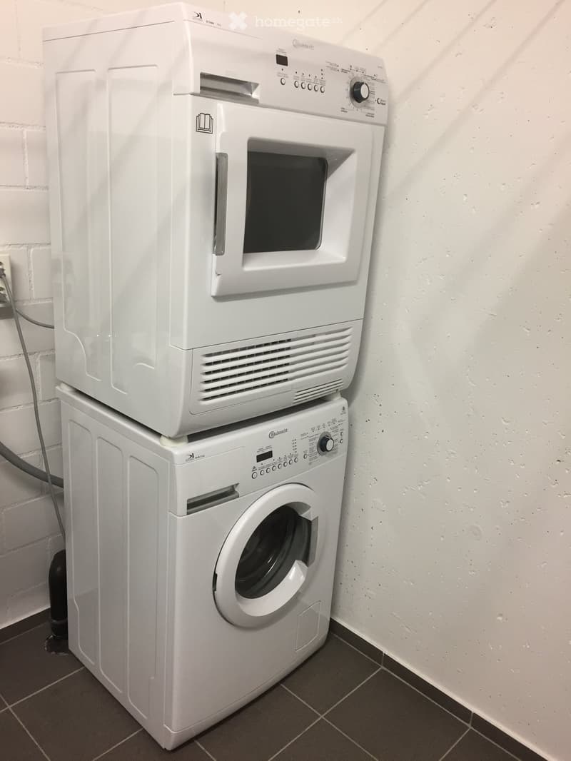 A compact bathroom with a white washing machine and a white dishwasher stacked on top of it, placed against a white wall with a brick accent.