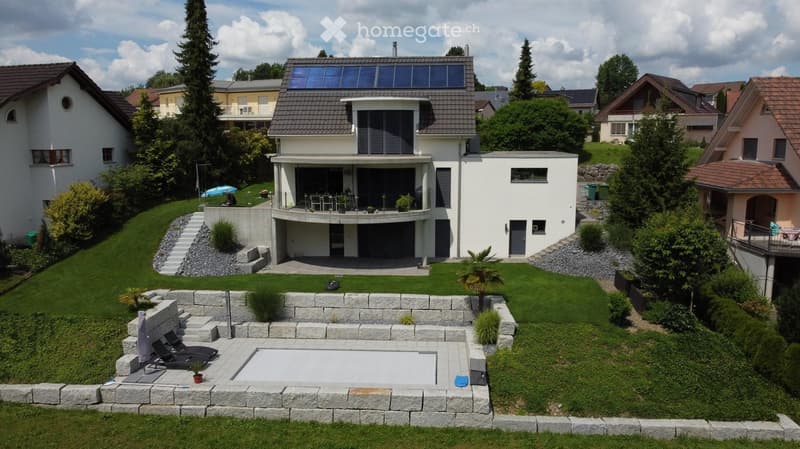 A modern two-story house with a large balcony, surrounded by a well-maintained garden with a swimming pool, lounge chairs, and various plants.