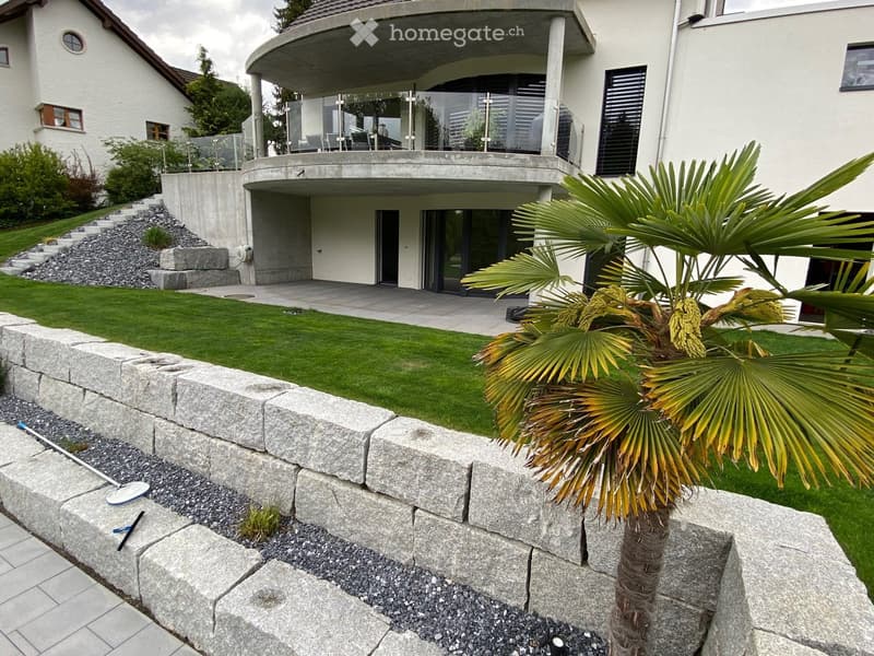 Modern house with a raised ground floor, a well-maintained lawn, a stone retaining wall, and a palm tree in a planter. The house features large windows and a balcony area with outdoor furniture.