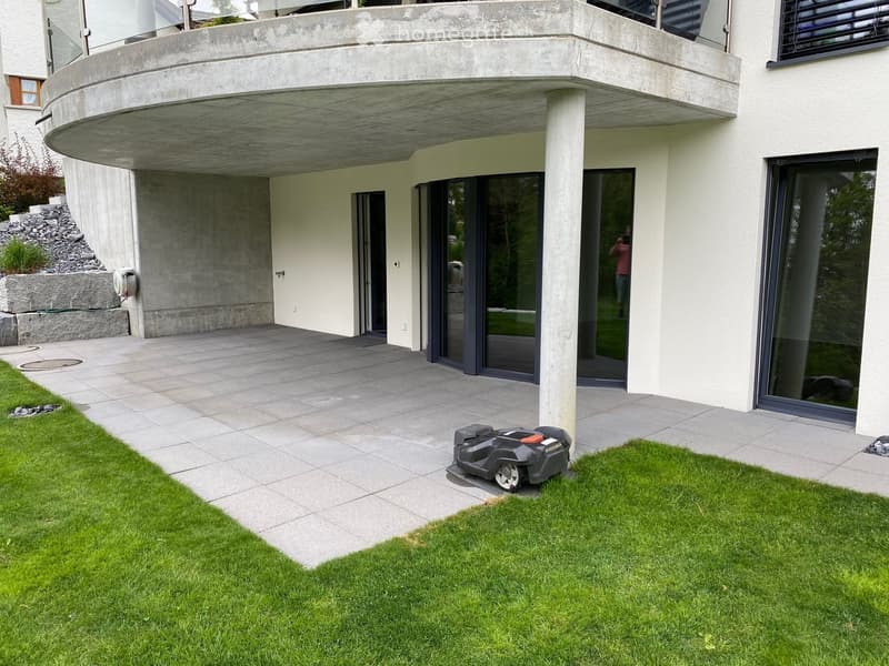 Modern house exterior with a raised ground floor, large windows, and a well-maintained lawn with a robotic lawn mower.