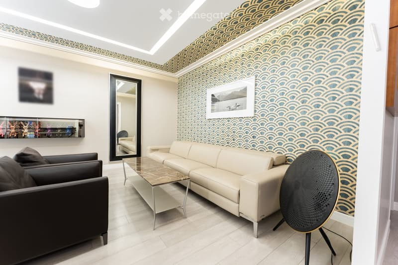 Modern living room with a white sectional sofa, a black leather chair, a glass coffee table, a large mirror, and a decorative fan. The walls feature a blue and gold patterned design, and the floor is tiled.