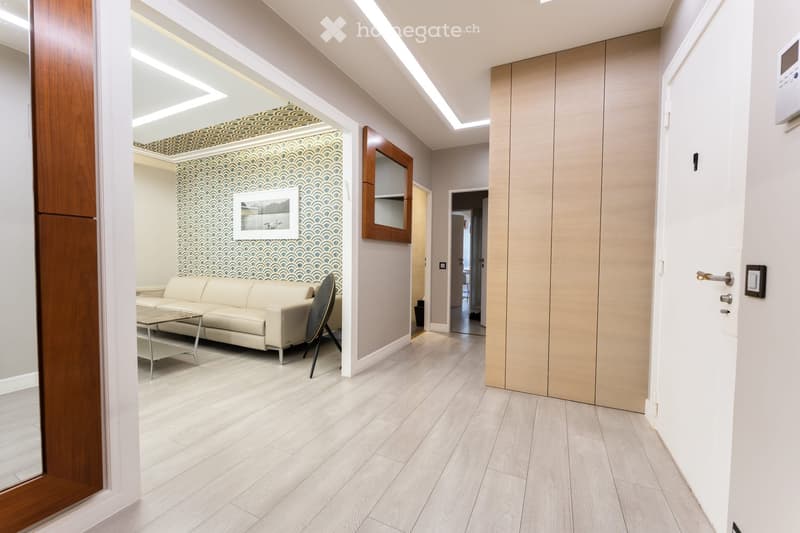 Modern living room with light-colored wooden flooring, a cream-colored sofa, a small coffee table, a modern chair, and a decorative wall with a framed picture. The room features sleek, contemporary design elements and recessed lighting.