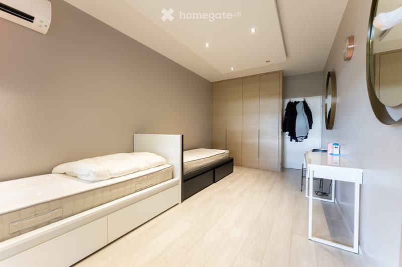 Modern bedroom with a single bed, built-in wardrobe, desk, and chair. The room features light-colored walls, wooden flooring, and recessed lighting.