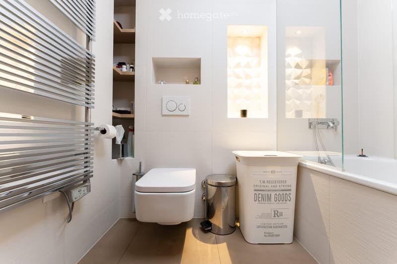 Modern bathroom with white tiles, a toilet, a bathtub with a glass enclosure, a towel rack, shelves, and a trash can.