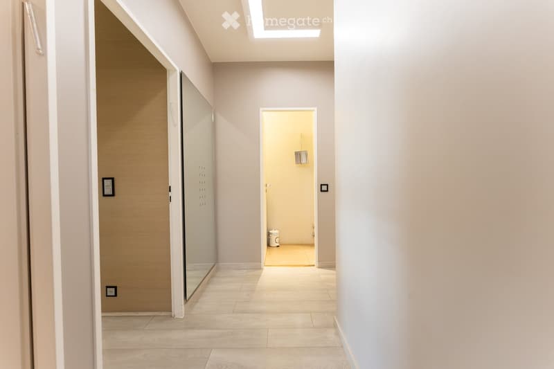 Modern hallway with light-colored tiles, white walls, and a glass door leading to another room. Features recessed lighting and a small trash bin in the adjacent room.