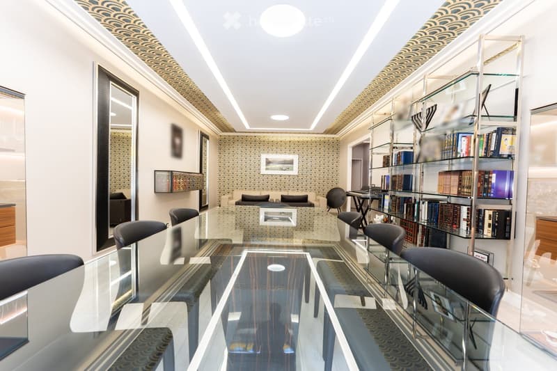 Modern office or meeting room with glass table, black chairs, bookshelf filled with books, large mirror, and decorative wall art.
