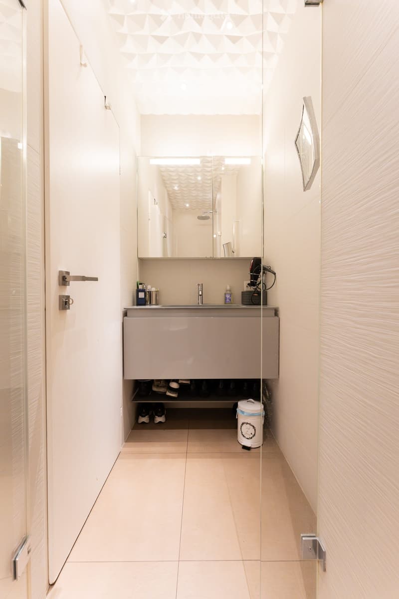 Modern bathroom with white tiles, a large mirror, a sink with a cabinet below, and a hair dryer on the counter. The floor is tiled, and there is a small trash bin near the cabinet.