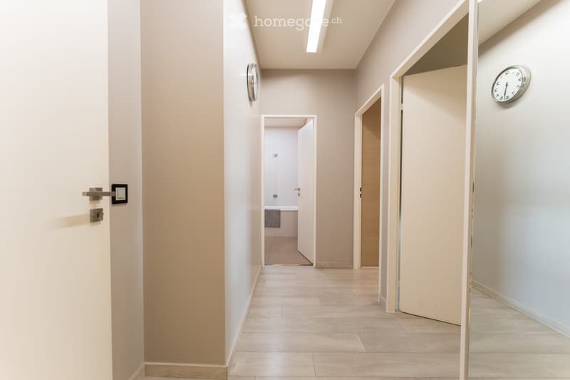 A clean, modern hallway with light-colored walls and tiled flooring, featuring two doors and a visible bathroom with a towel hanging on the wall.