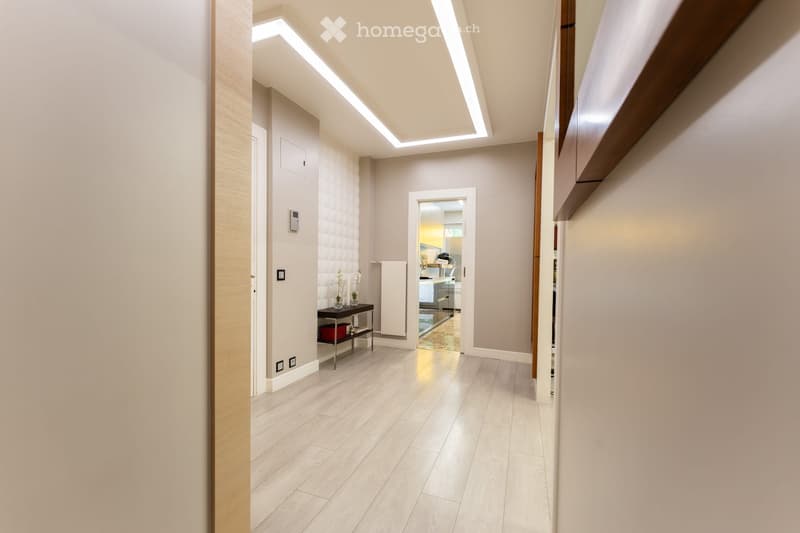 Modern hallway with light wooden flooring, white walls, and recessed ceiling lighting. Features a small black console table with vases and a radiator cover, leading to a kitchen area visible in the background.