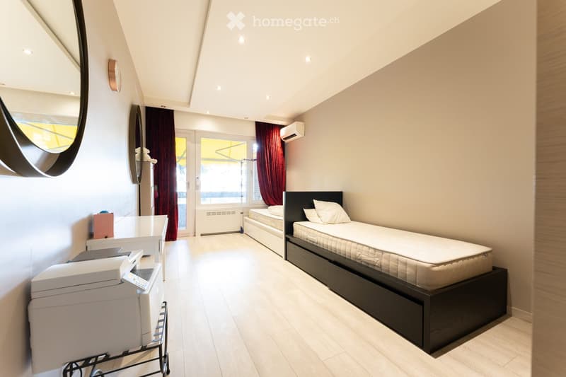 A modern, well-lit bedroom with two single beds, a nightstand, a mirror, a printer, and a small table with a chair.