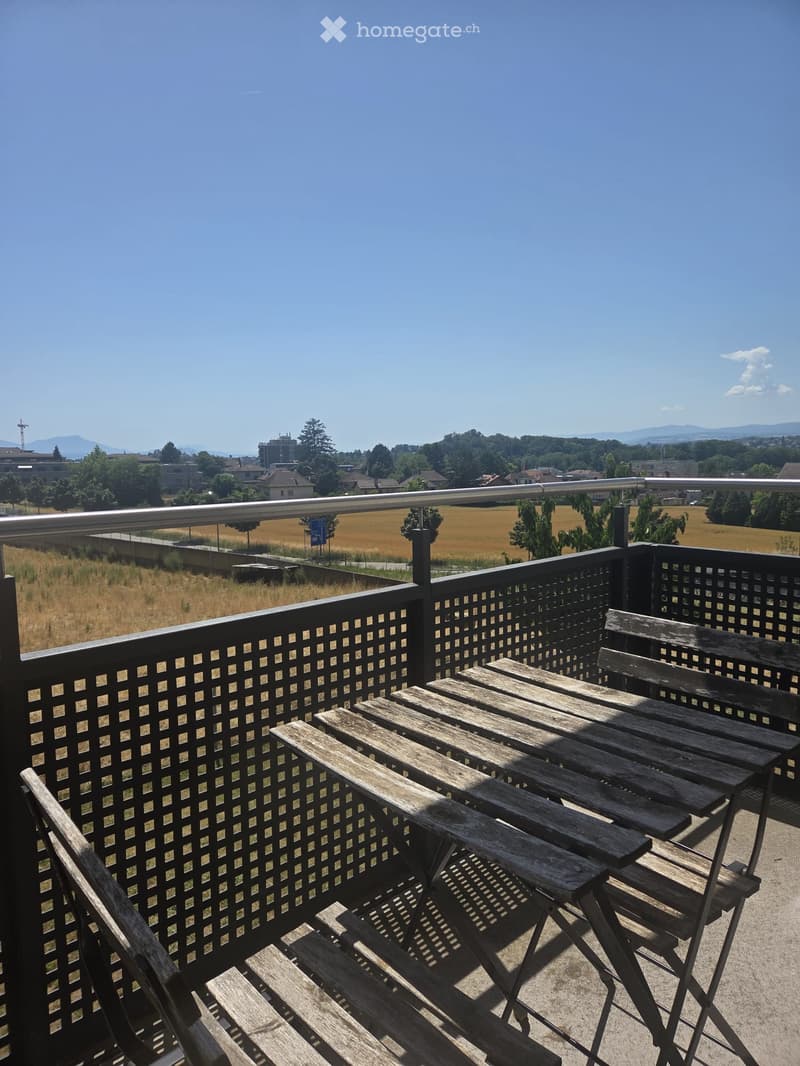 Balcony with wooden table and chairs, overlooking a rural landscape with fields and distant buildings.