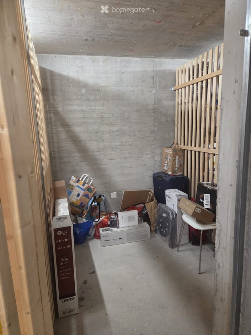 A small, unfinished room with exposed concrete walls and floor, containing a white plastic chair, a black suitcase, several cardboard boxes, and a small stool.
