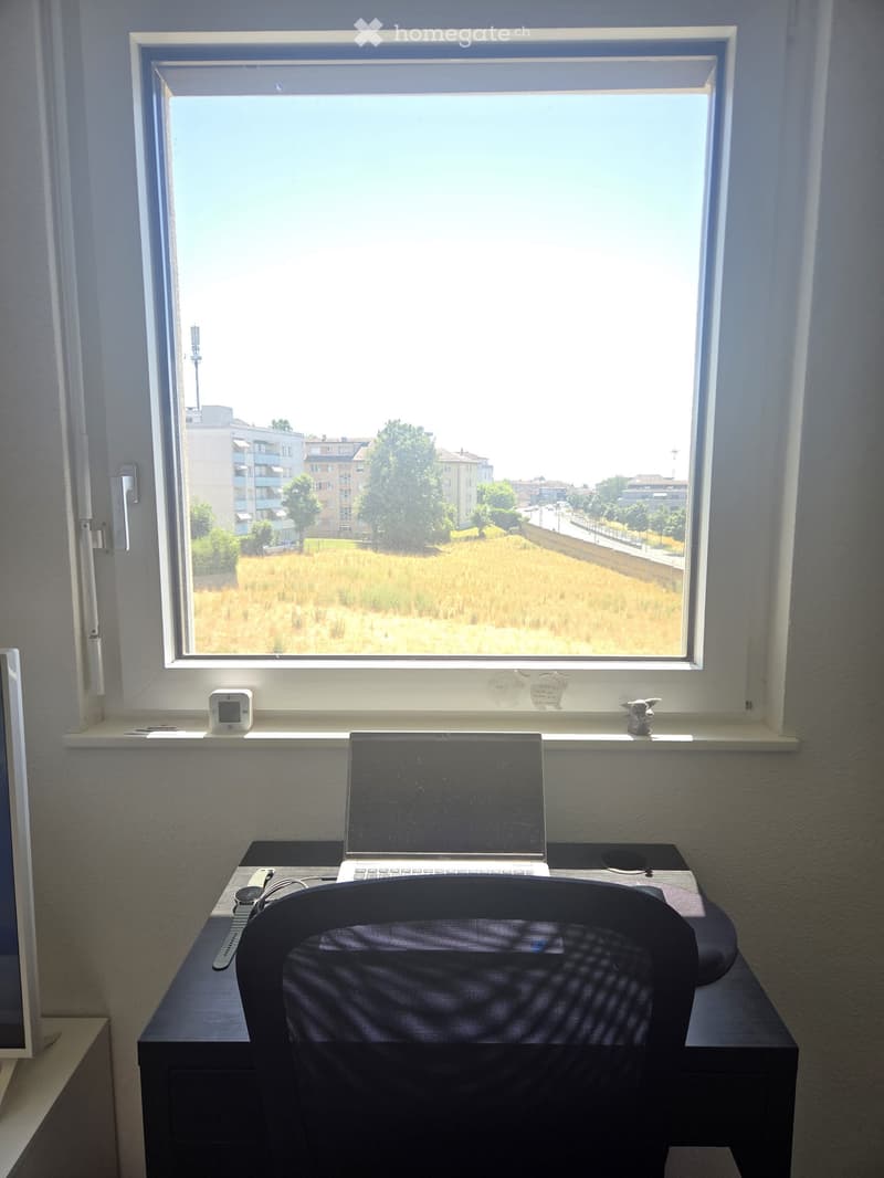 A furnished room with a desk, laptop, chair, and a large window overlooking a field and buildings.