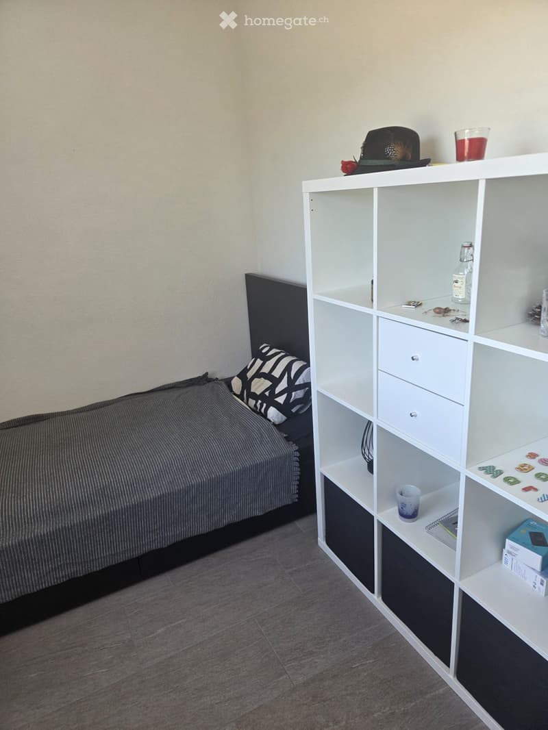 A small bedroom with a single bed, a modern white shelving unit, and various personal items on the shelves.
