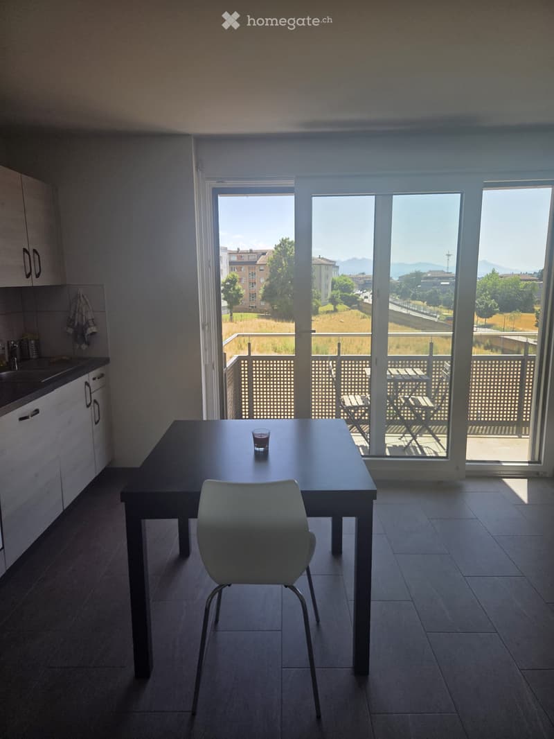 Modern kitchen-dining area with a dark wooden dining table, white chair, and a cup on the table. Large windows and sliding doors open to a balcony with outdoor furniture and a scenic view of the city and mountains.