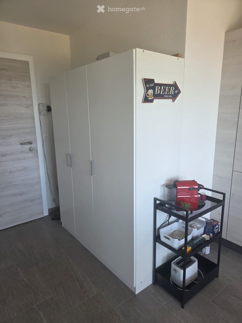 A small, modern kitchen area with a refrigerator, a cart holding a coffee maker, toaster, and other small appliances, and a sign that reads "Ice Cold BEER Served Here."