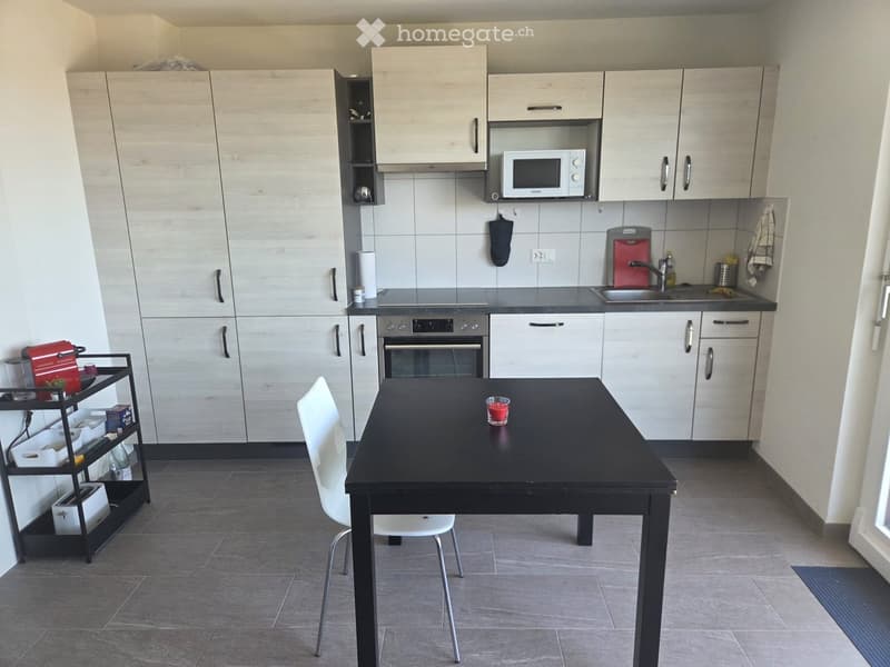 Modern kitchen with light wood cabinets, stainless steel appliances, a black dining table with chairs, and a small cart with items.