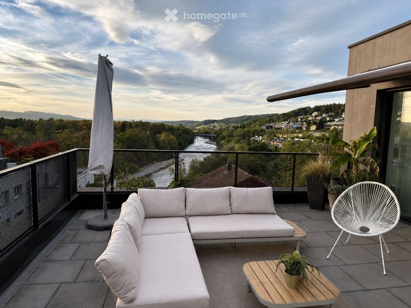 Modern balcony with a white sectional sofa, a white wicker chair, a small wooden table with a potted plant, and an umbrella, overlooking a scenic city view.