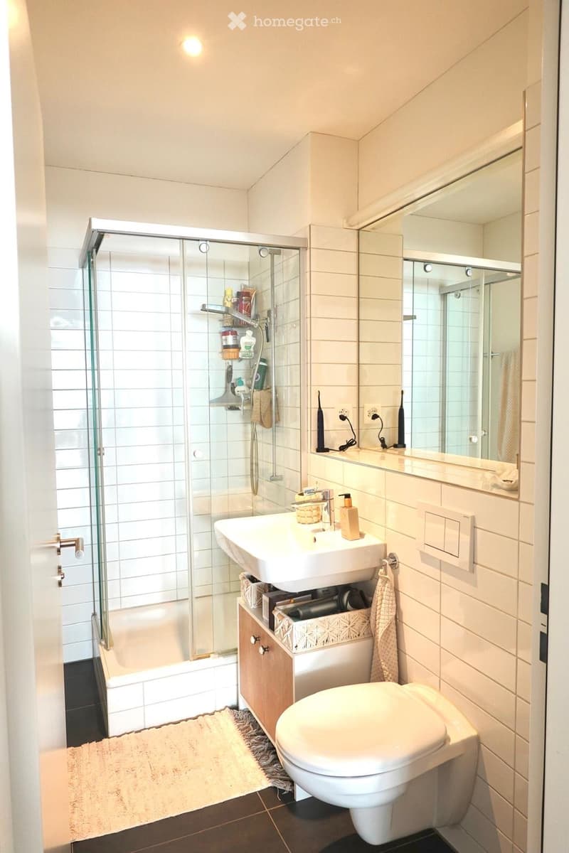 Modern bathroom with white tiles, a glass-enclosed shower, a sink with a cabinet, a toilet, and a large mirror above the sink. The space includes a towel and various bathroom accessories on the sink and shower shelf.