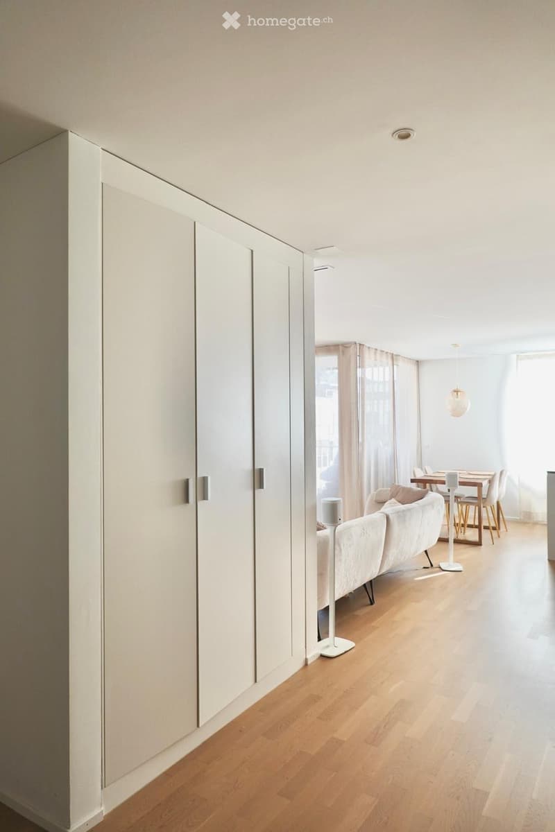 Open-plan living and dining area with light wooden flooring, white walls, and modern furniture including a sofa, dining table, chairs, and lamps.