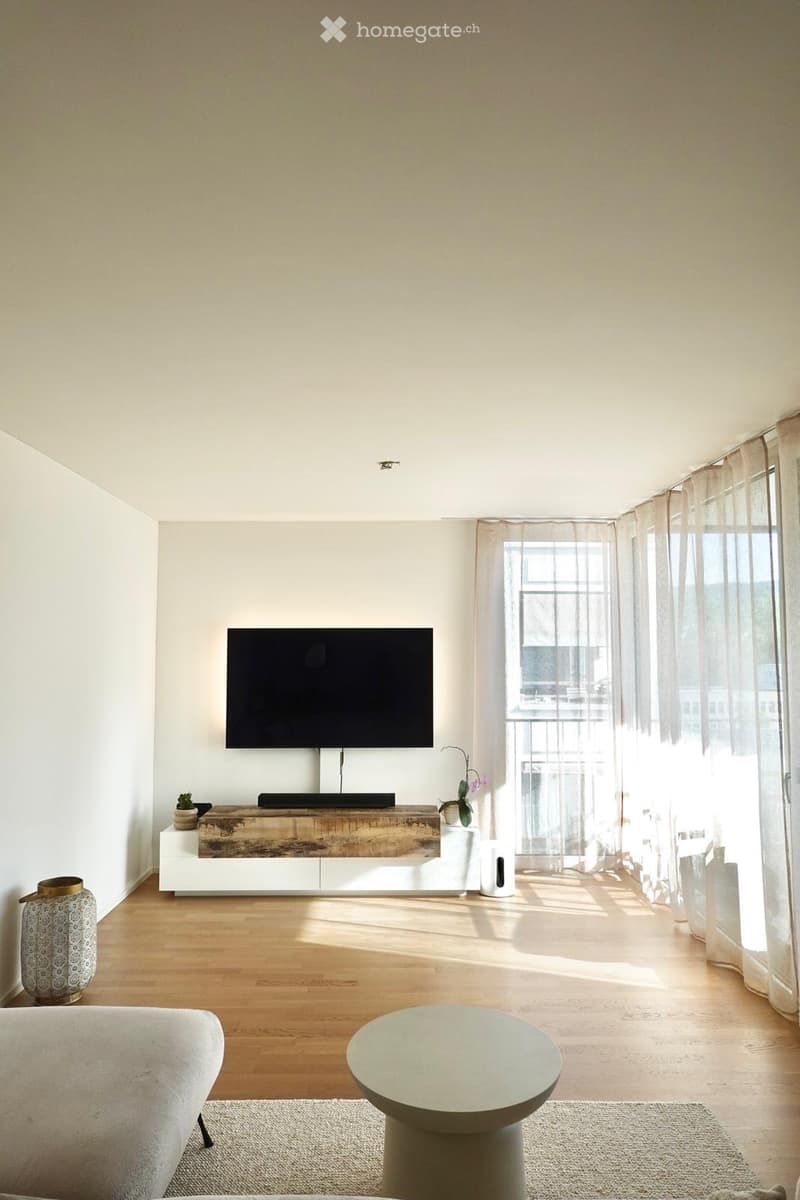 Modern living room with light wooden flooring, white walls, large windows with sheer curtains, a mounted TV, a white entertainment unit, a round coffee table, and a grey sofa.