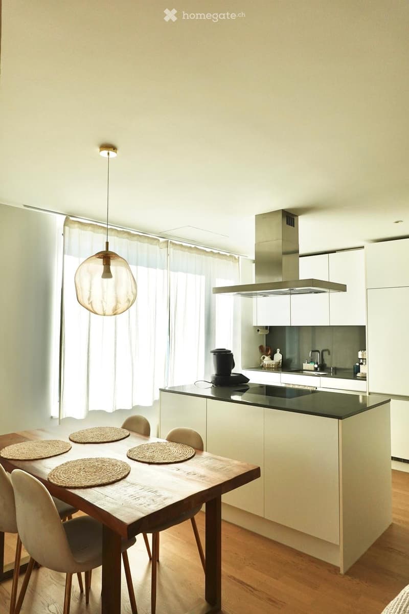 Modern kitchen with white cabinets, a wooden dining table with four chairs, and a black countertop. A coffee maker sits on the counter, and a range hood is visible above the sink.