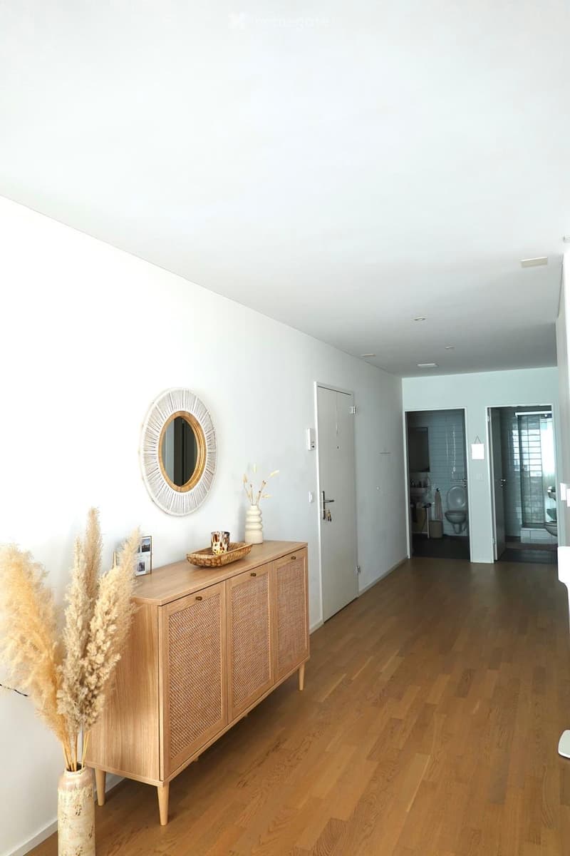 A modern hallway with light wooden flooring, white walls, and a decorative sideboard with a mirror, vase, and tray. The hallway leads to a bathroom with visible fixtures.