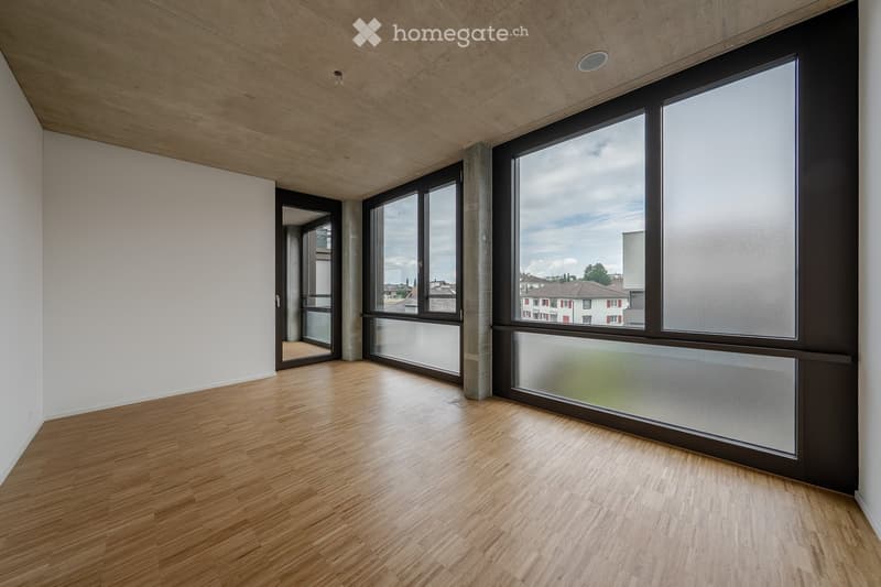 Empty room with wooden flooring, white walls, large windows, and a door leading to another space.