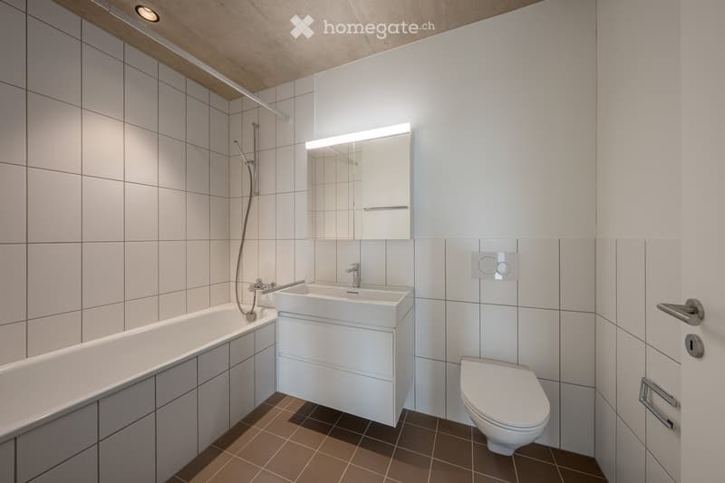 Modern bathroom with white tiles, a bathtub, a sink with a modern faucet, a toilet, and a large mirror with built-in lighting. The floor is tiled in a warm brown color.