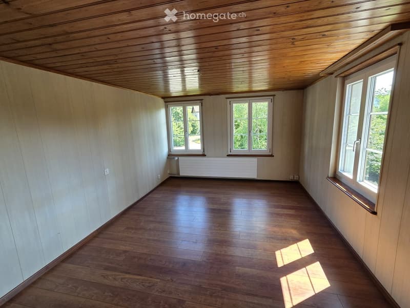 Empty room with wooden flooring, white walls, and multiple windows allowing natural light to enter. The room features a wooden ceiling and a radiator under one of the windows.