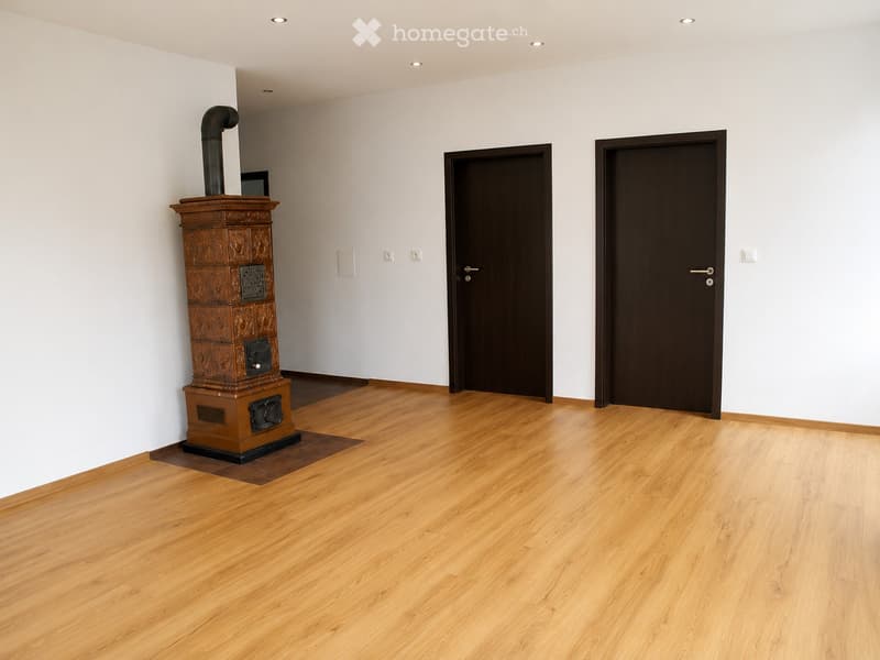 Empty room with wooden flooring, white walls, and a decorative wooden stove with a chimney. Two dark wooden doors are visible.